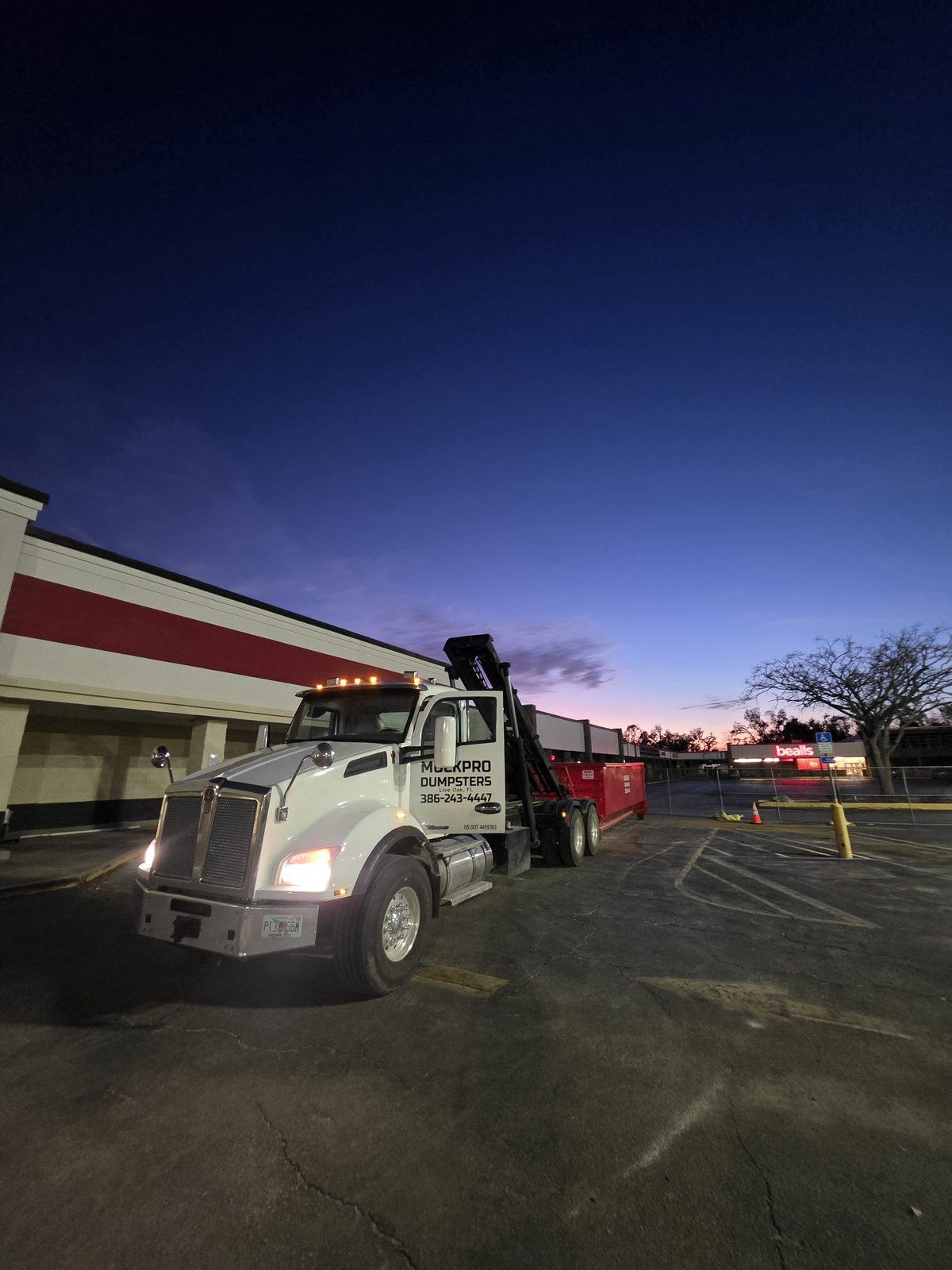 Muckpro roll-off truck delivering a dumpster at a commercial plaza at dusk